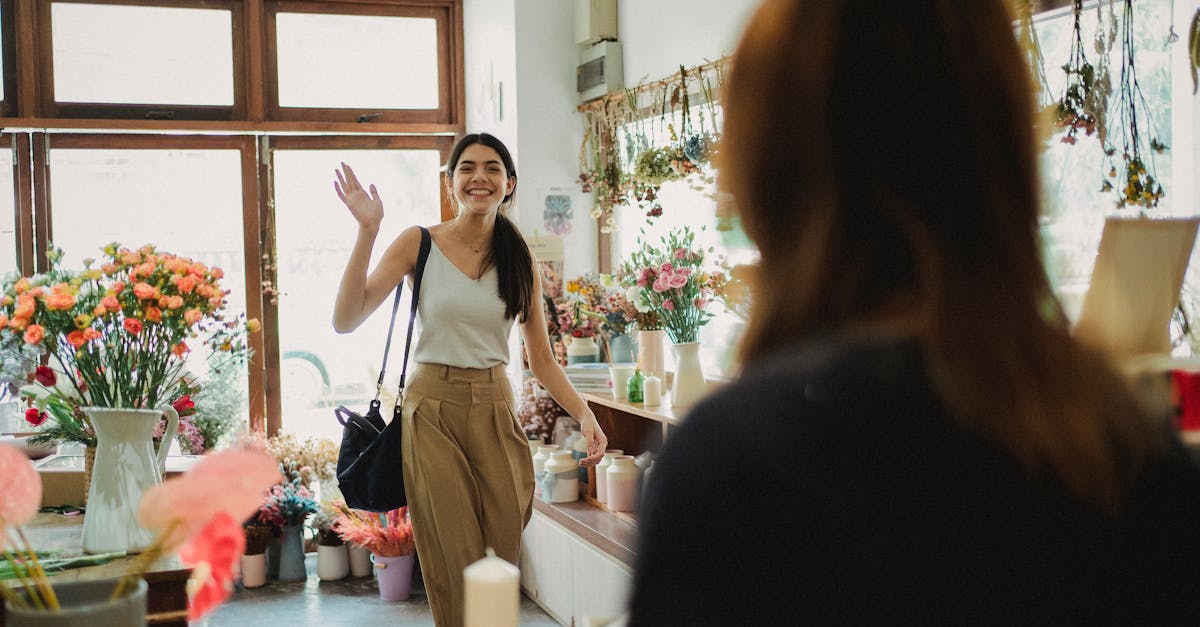 Cheerful lady with dark hair in casual clothes coming to flowers shop and waving to blurred florist in daytime