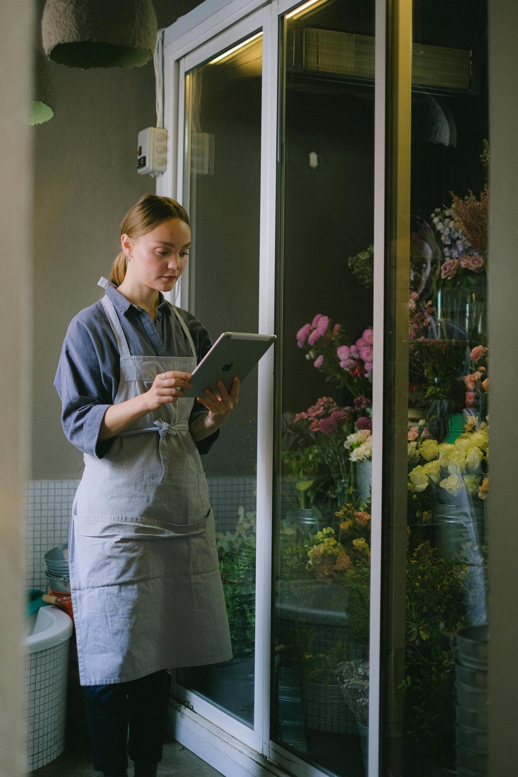 A woman florist in an apron uses a tablet in her flower shop, surrounded by colorful flowers.